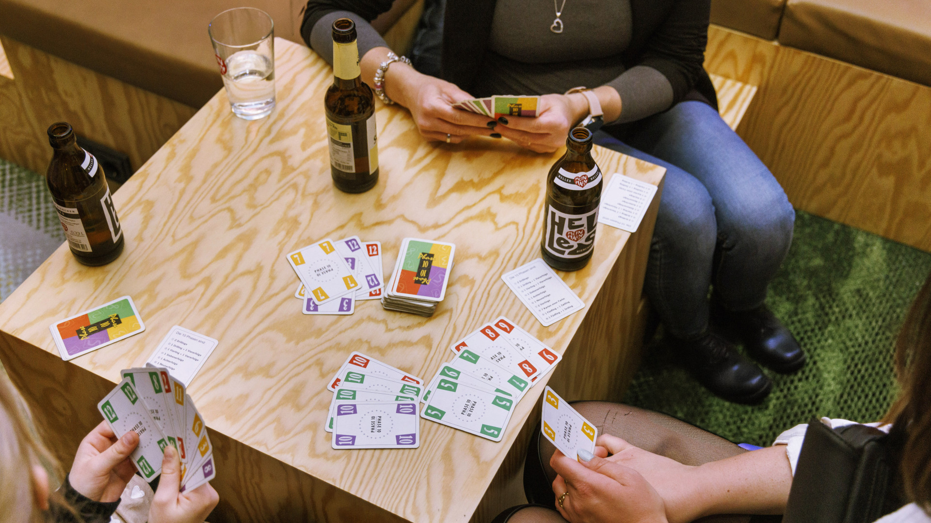 Mehrere Hotelgäste spielen Karten um einen kleinen Holztisch mit verschiedenen Getränken darauf. 