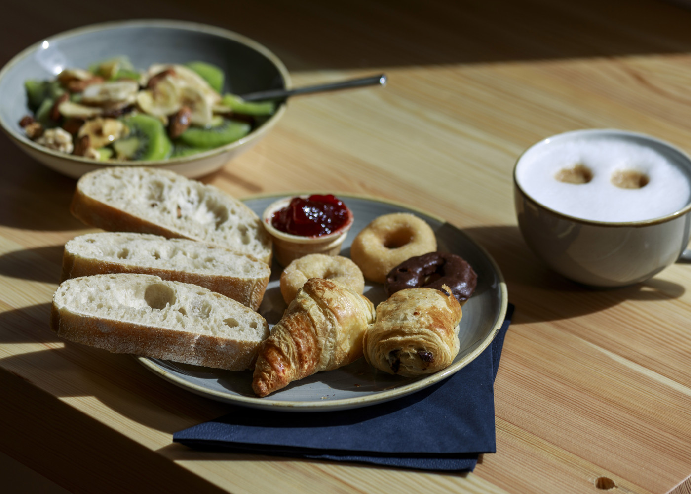 Frühstücksteller mit Croissant, Baguette, Marmelade und Gebäck, daneben Cappuccino und Müsli mit Obst.