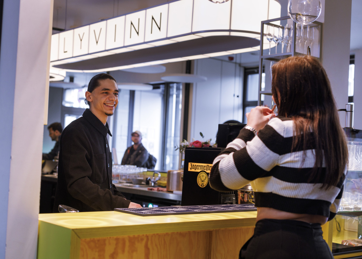 Mitarbeiter hinter der Hotelbar unterhält sich freundlich mit einer Frau im gestreiften Oberteil.