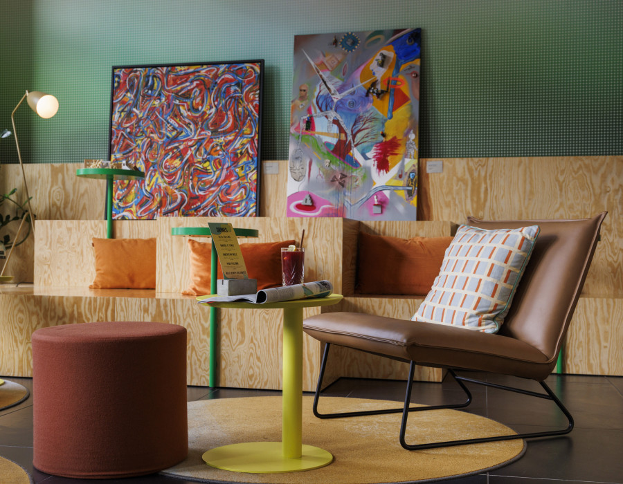 Stilvolle Lounge-Ecke mit farbenfrohen Bildern, Holzmöbeln, Kissen und Getränken auf kleinen Tischen.