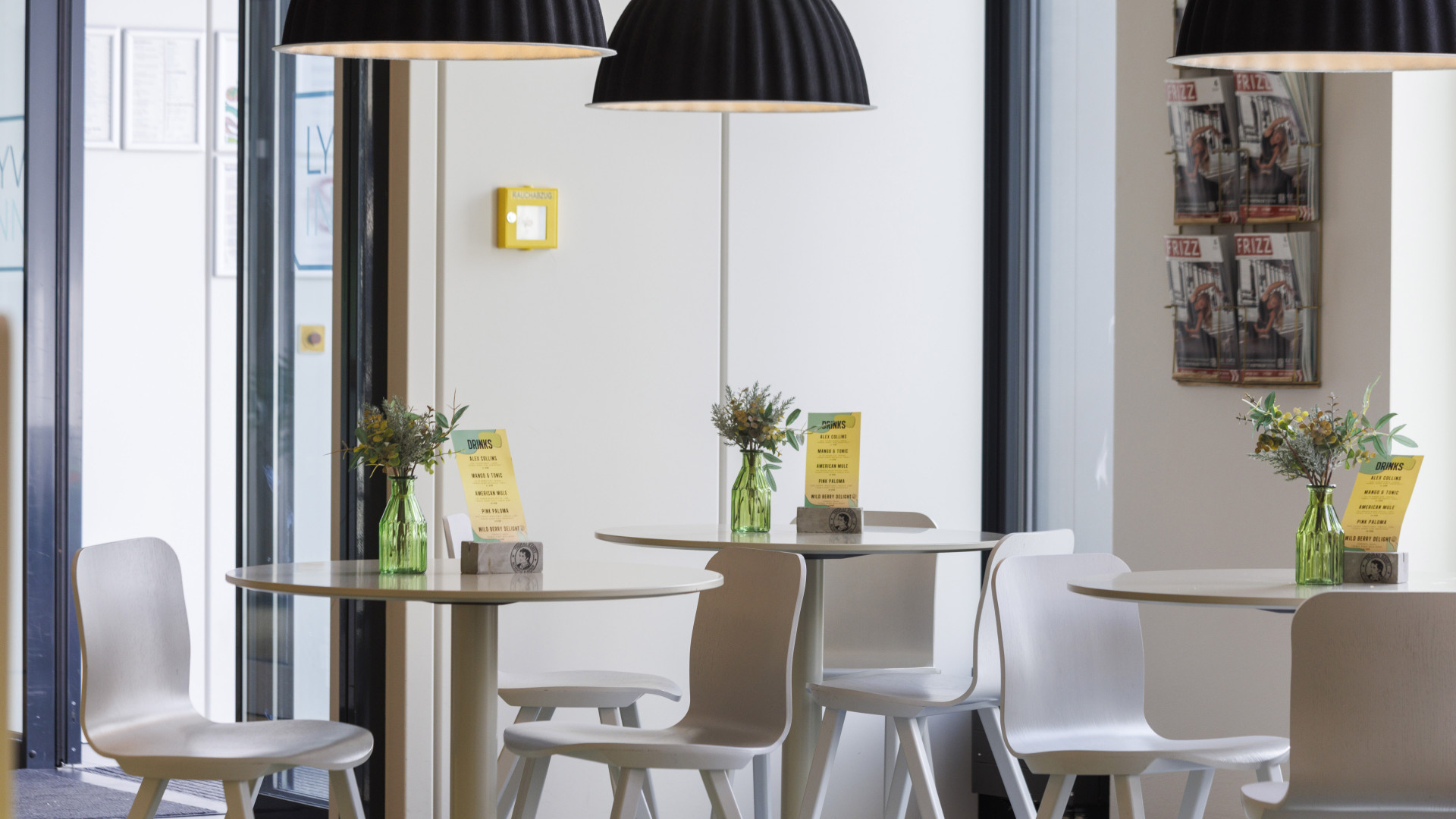 Heller Café- Bereich in einer Hotellobby mit weißen Stühlen, Tischen und dunklen modernen Lampen über den Tischen.