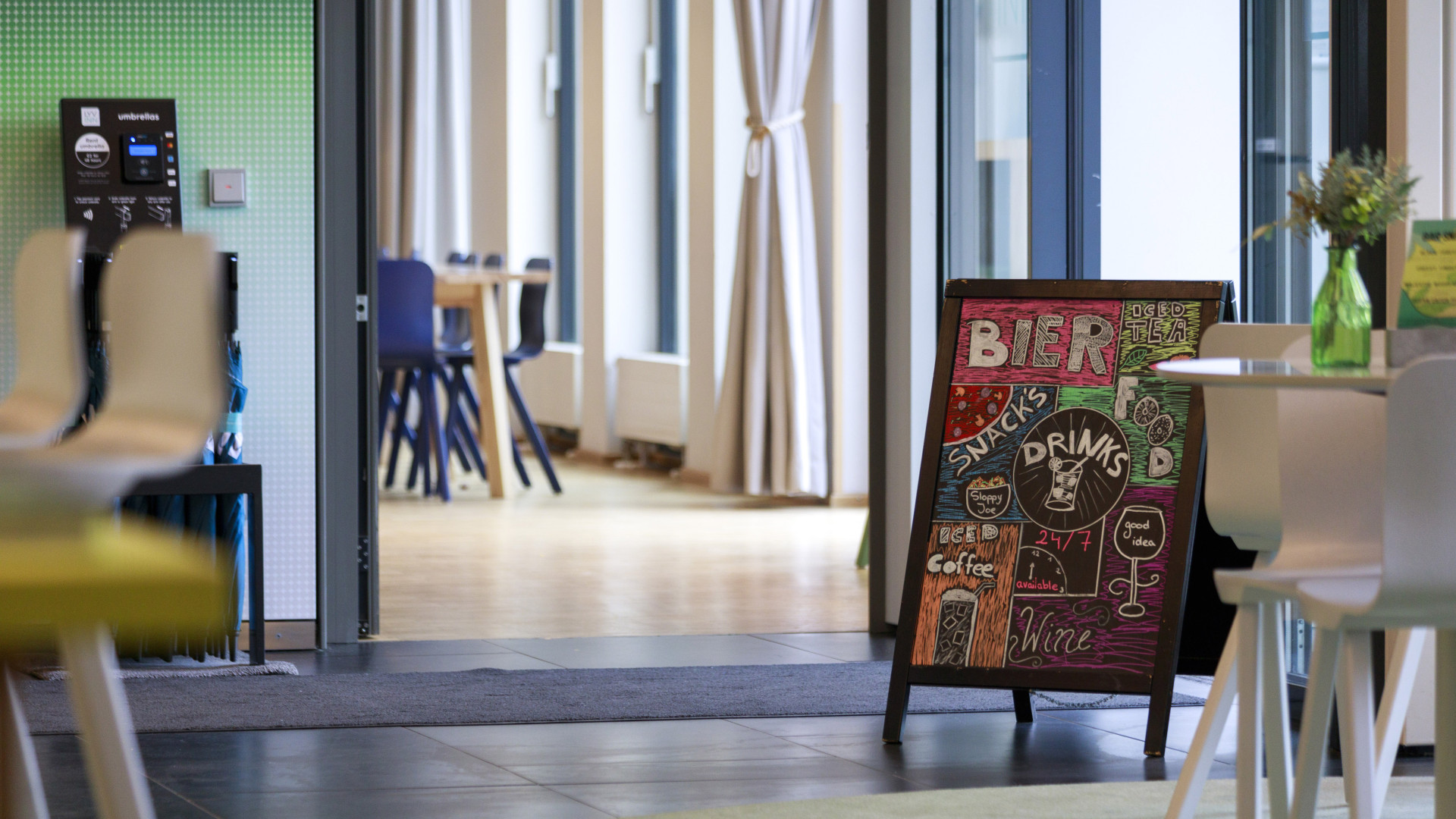 Moderner Eingangsbereich eines Hotels mit Tischen, Sitzecke und einem Aufsteller, der die Hotelbar bewirbt.