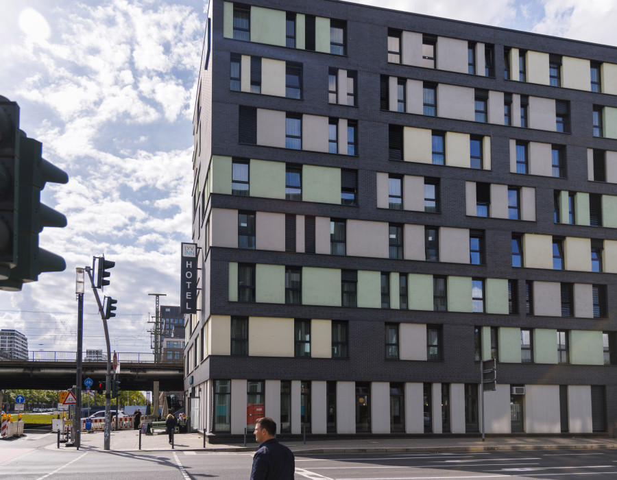 Modernes Hotelgebäude an einer Straßenecke mit vielen Fenstern, Fußgängern und Ampelanlage davor.