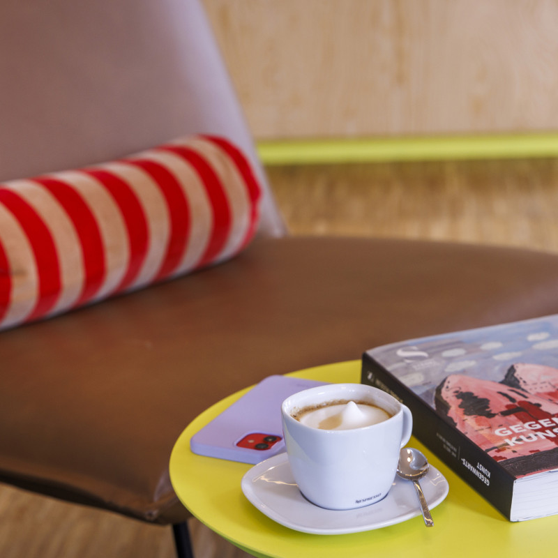 Eine Tasse Kaffee auf einem runden Beistelltisch, daneben ein brauner Stuhl mit einem roten, runden Kissen