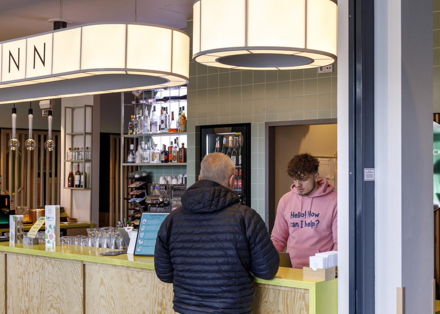 Gast spricht an der Bar mit einem Mitarbeiter im pinken Hoodie, im Hintergrund Flaschenregal und Beleuchtung.