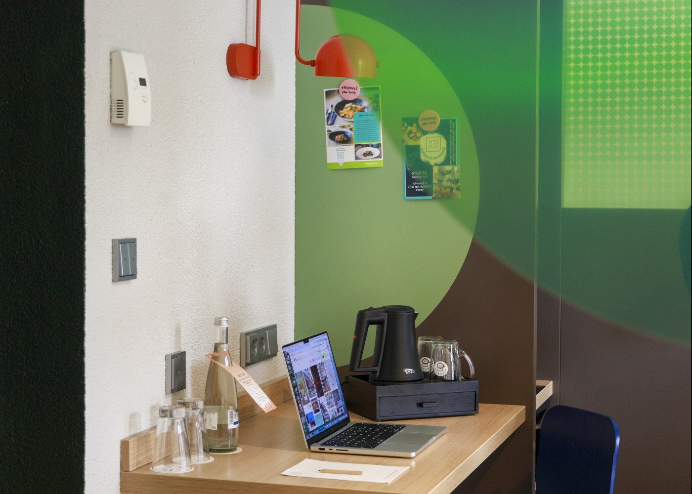 Schreibtisch mit Wasserkocher, Gläsern und Wandkunst, daneben Laptop und auffällige rote Wandlampe.