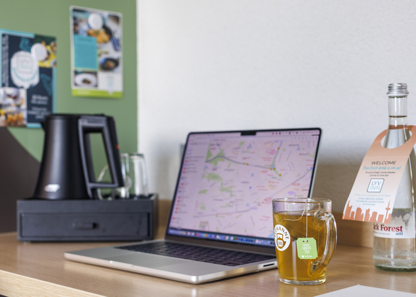 Aufgeräumter Schreibtisch mit Laptop, Teeglas, Wasserflasche und Kaffeemaschine vor Infowand im Hotelzimmer.