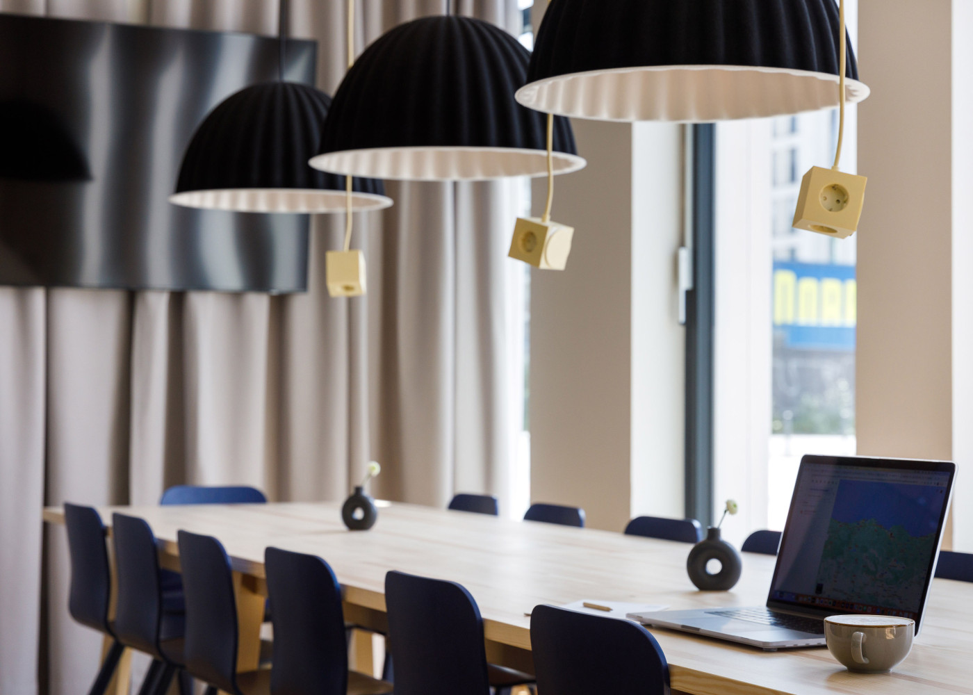 Langer Besprechungstisch mit Laptop, Hockern und modernen schwarzen Hängelampen in lichtdurchflutetem Raum.