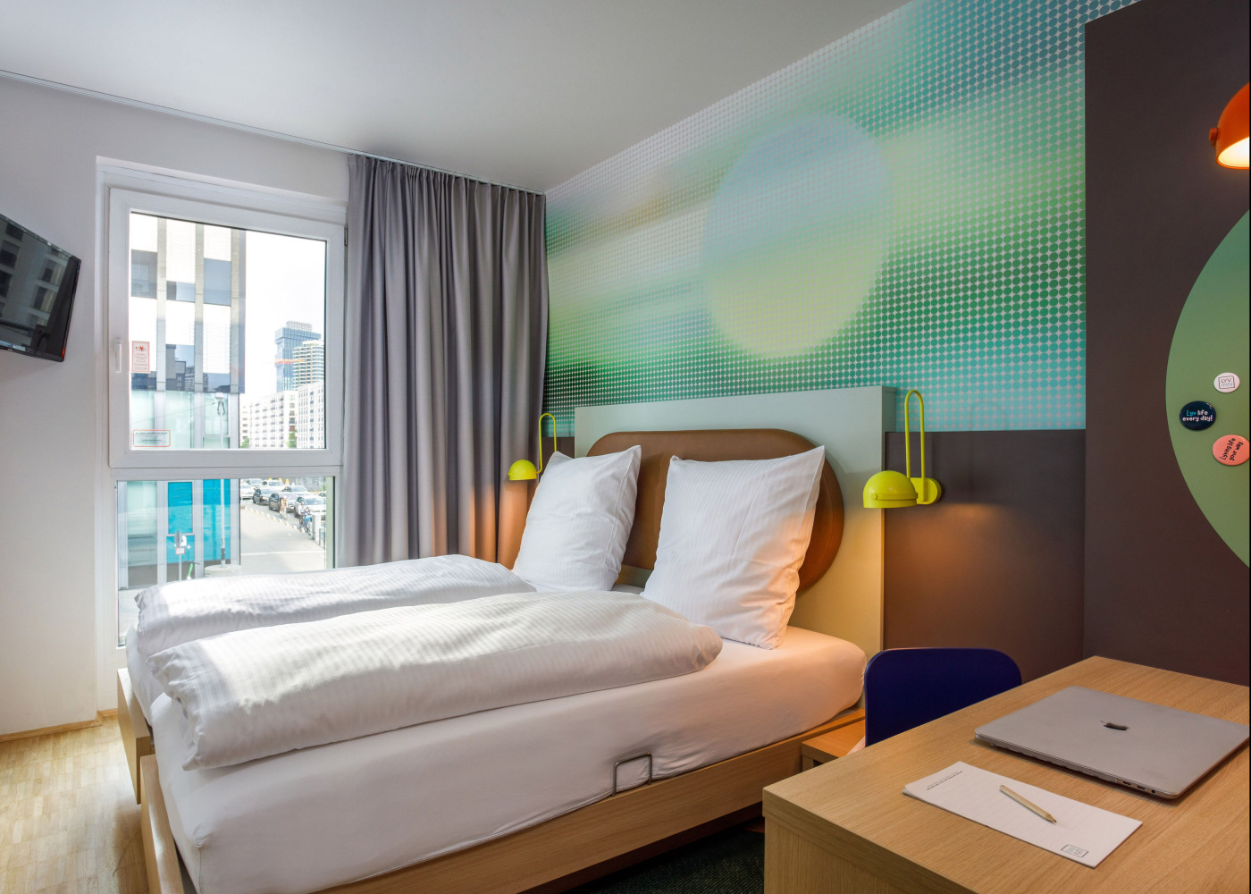 Modernes Hotelzimmer mit Doppelbett, gelben Wandlampen, abstrakter Wandgestaltung und Fensterblick.