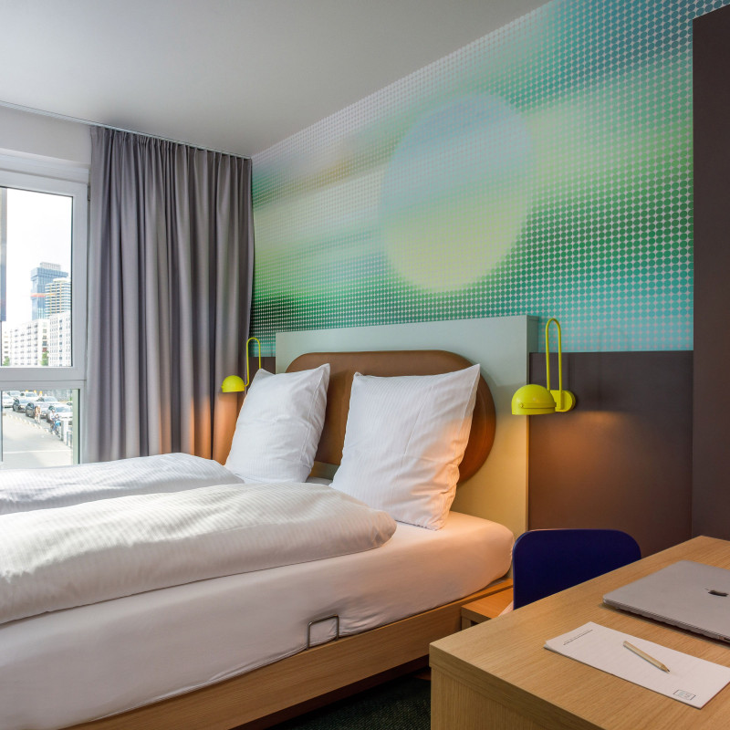 Helles Hotelzimmer mit Doppelbett, farbiger Wandgestaltung, großem Fenster und Schreibtisch im Vordergrund.