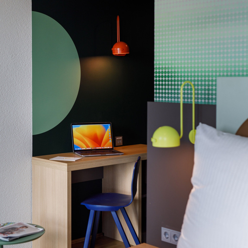 Kompakter Schreibtisch mit Laptop und roter Lampe neben einem Bett, Wandgestaltung mit grünem Kreis.