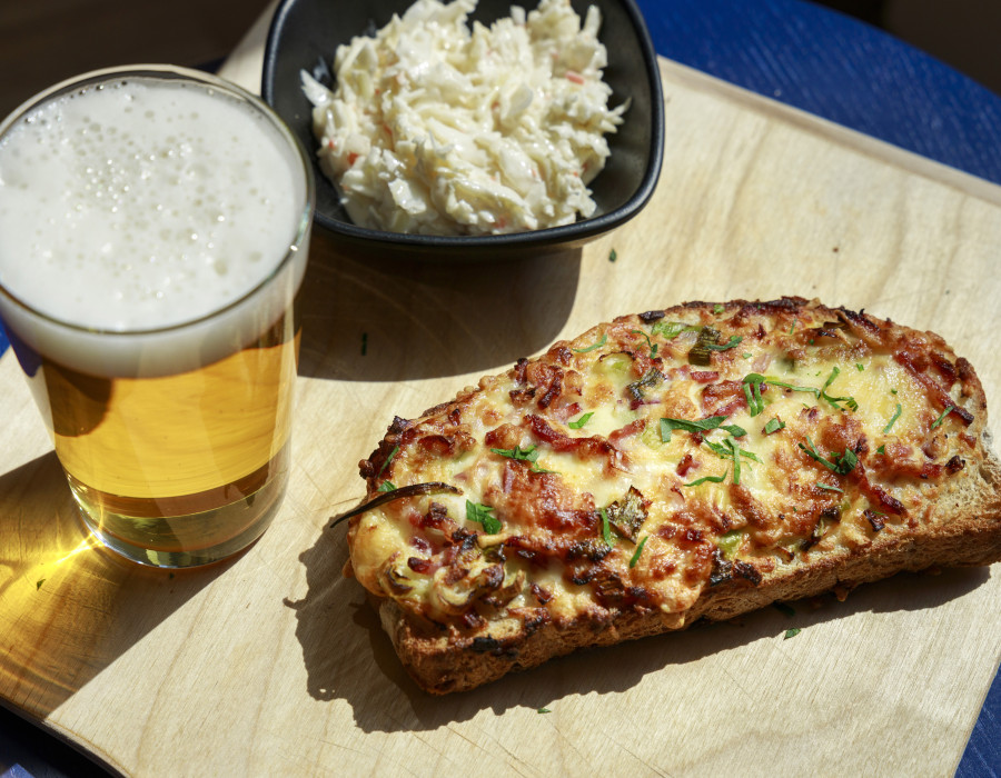 Überbackenes Käsebrot auf Holzbrett mit Glas Bier und Krautsalat in Schale bei Tageslicht serviert.