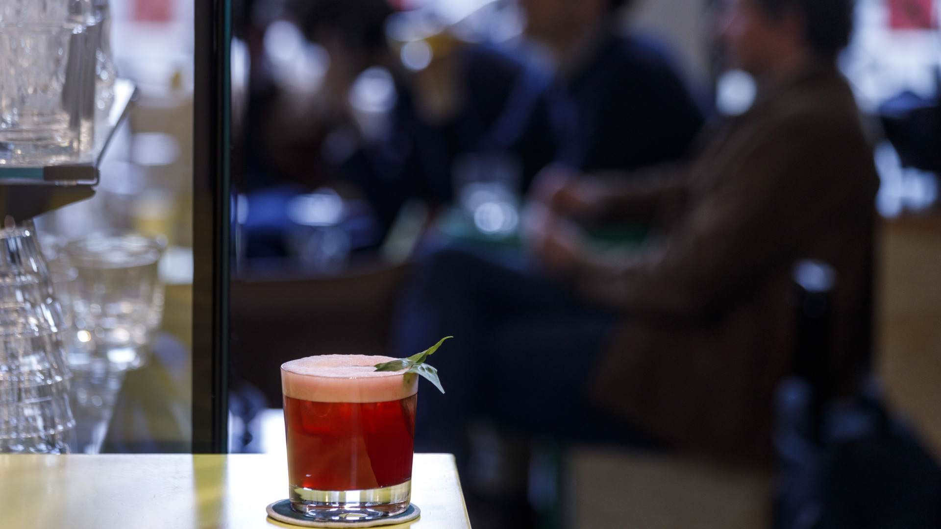 Ein roter Cocktail in einem schönen Glas steht auf dem Tresen einer modernen Hotelbar, Menschen im Hintergrund.