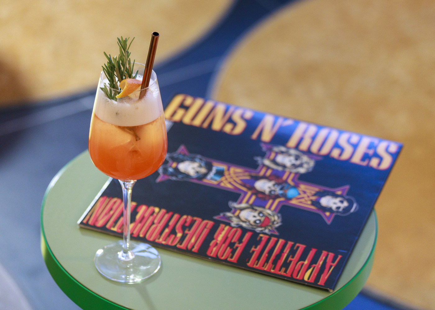 Cocktailglas mit Deko auf grünem Tisch, darunter ein Guns N’ Roses-Albumcover als Unterlage im Lounge-Bereich.