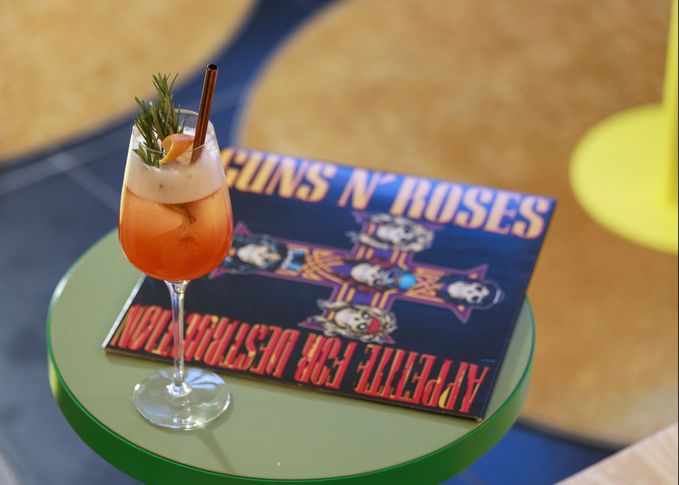 Cocktailglas mit Deko auf grünem Tisch, darunter ein Guns N’ Roses-Albumcover als Unterlage im Lounge-Bereich.