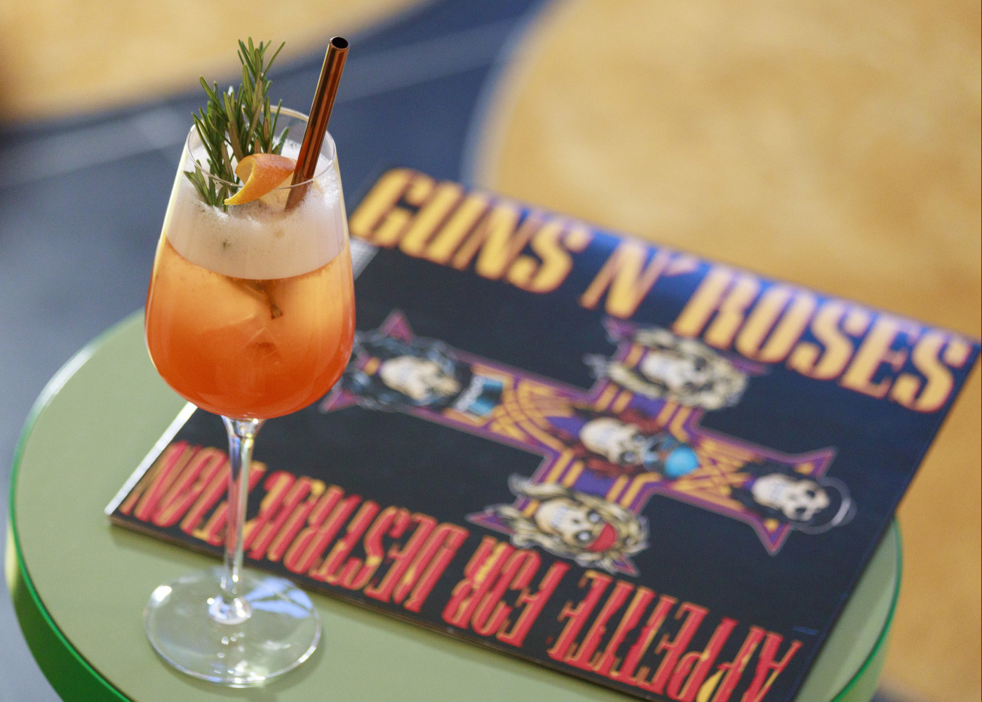 Auf einem grünen Rundtisch steht ein Cocktail in einem hohen Weinglas und eine Schallplatte von Guns N’ Roses.