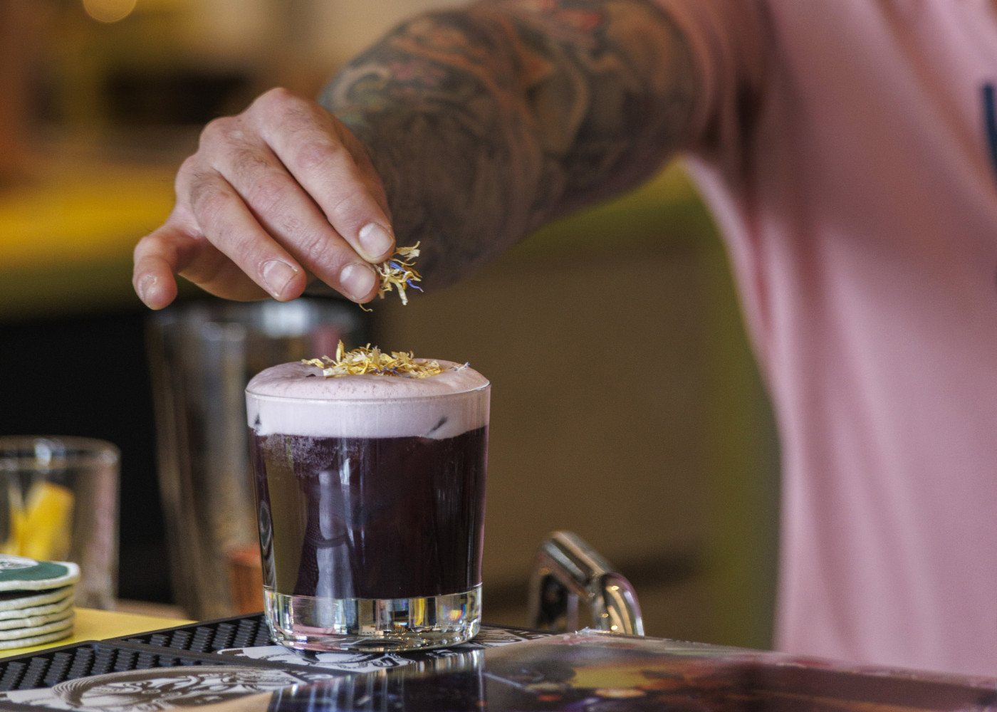 Tätowierte Hand garniert einen Cocktail mit violettem Schaum vorsichtig mit getrockneten Blüten als Deko.