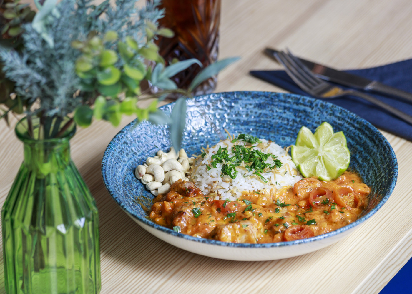 Teller mit Reis, Curry, Limettenspalten und Cashews auf Holztisch, daneben Vase mit frischen Blumen.