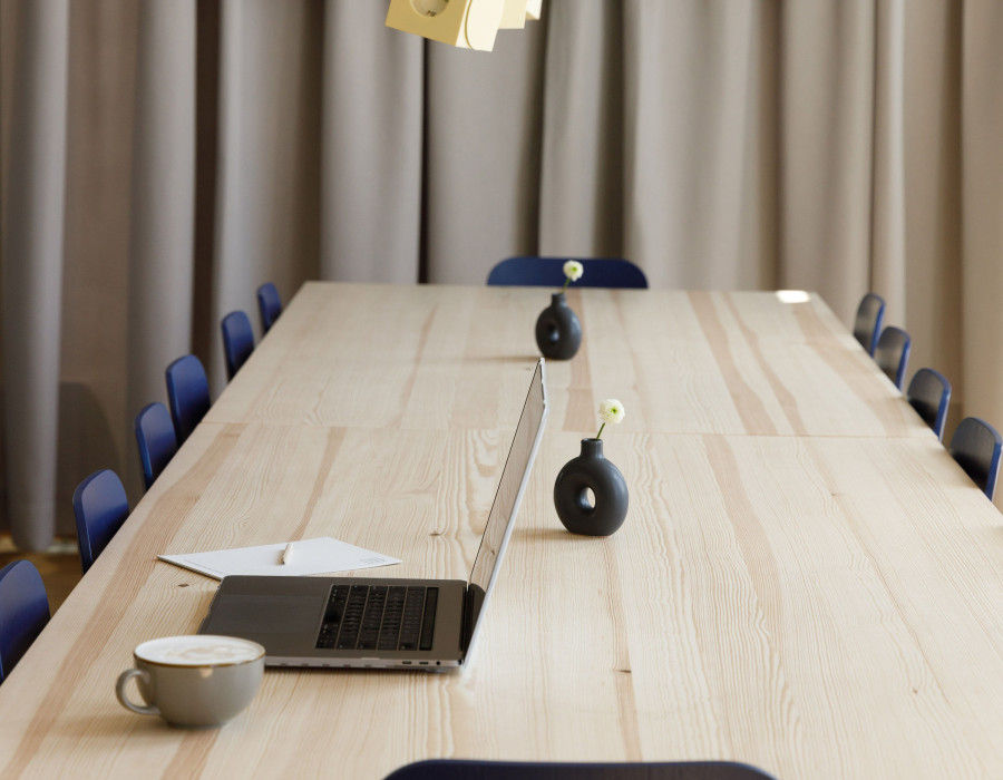 Großer Konferenztisch aus Holz mit aufgeklapptem Laptop, Kaffeetasse und Vorhängen im Hintergrund.
