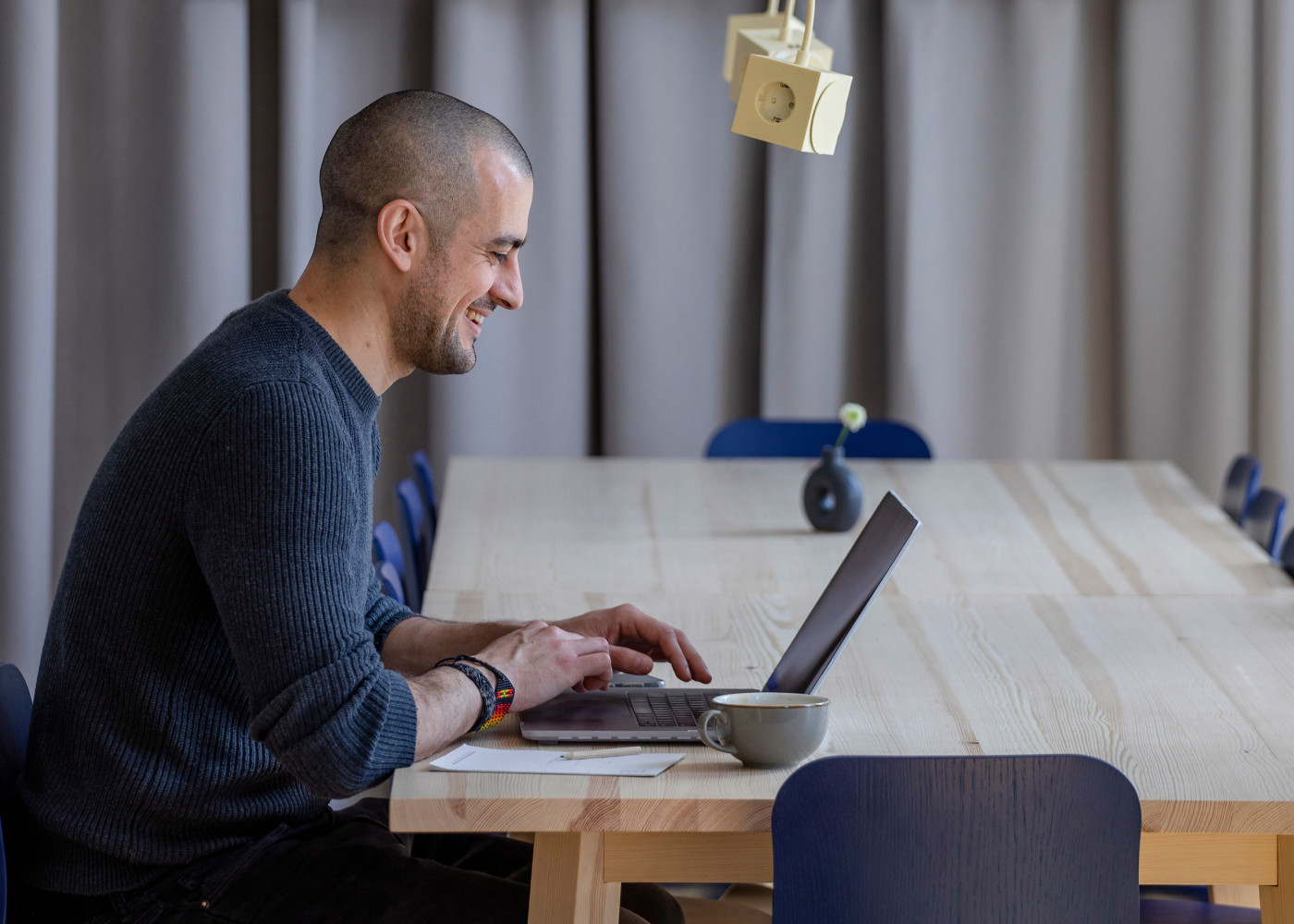 Lächelnder Mann arbeitet an einem Laptop an einem langen Holztisch mit Vorhängen und Kaffeetasse daneben.
