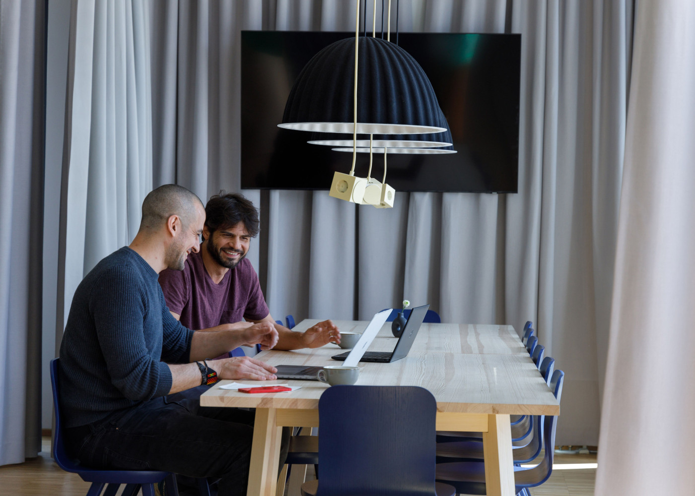 Zwei Männer arbeiten gemeinsam lachend an einem Laptop in einem modernen Raum mit Vorhängen und TV.