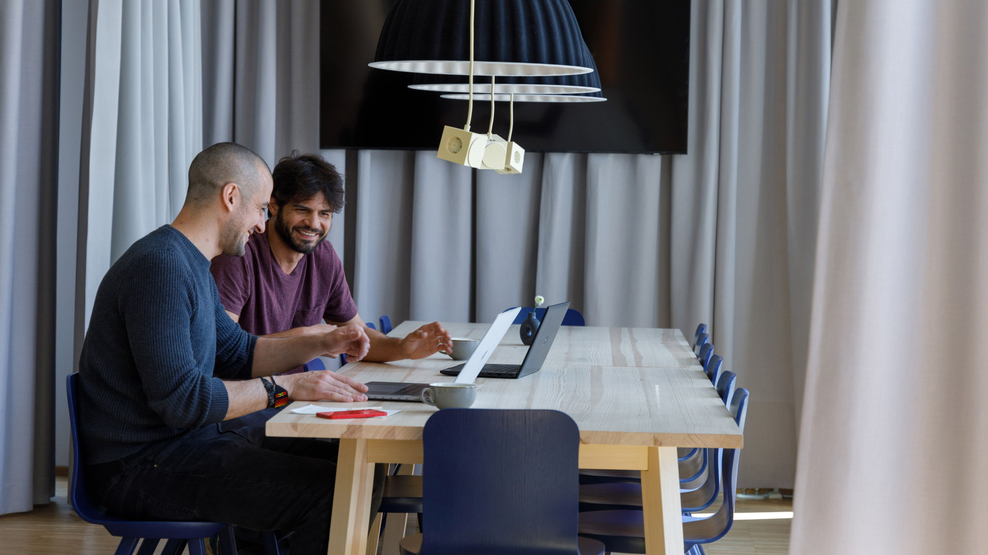 Zwei Männer sitzen nebeneinander an einem großen Tisch in einem Co-Working Bereich und besprechen sich.