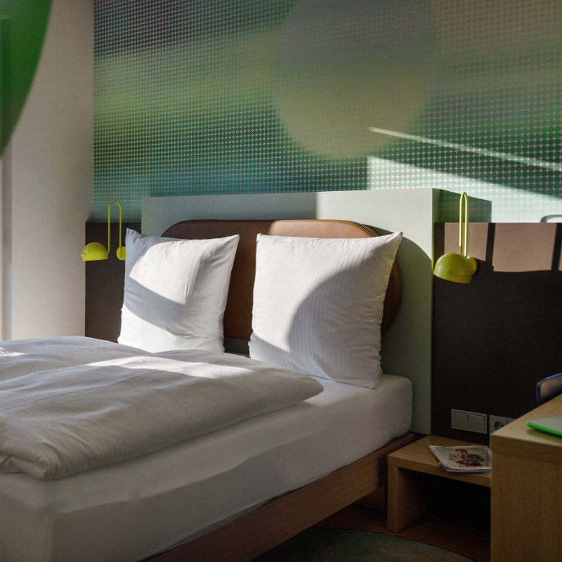 Helles Hotelzimmer mit großem Doppelbett, Nachttischen, Tageslicht und stilisiertem grünem Wanddesign.