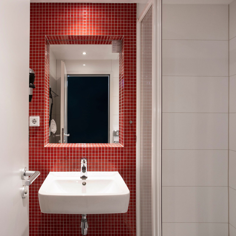 Modernes Bad mit weißem Waschbecken, großem Spiegel und auffälliger roter Wandfliese hinter dem Waschplatz.