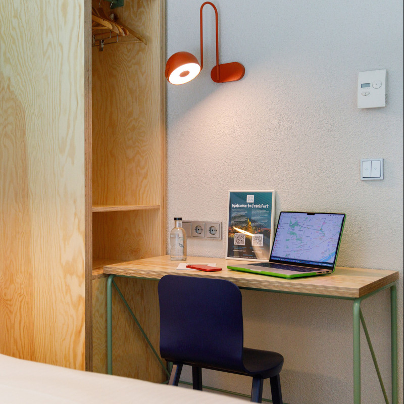 Moderner Holzschreibtisch mit Laptop und roter Lampe neben offenem Kleiderschrank und Bett im Hotelzimmer.