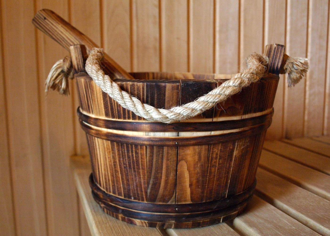 Holzeimer mit Seilgriffen und Schöpfkelle in einer Sauna, auf einer Holzbank vor vertäfelter Wand.