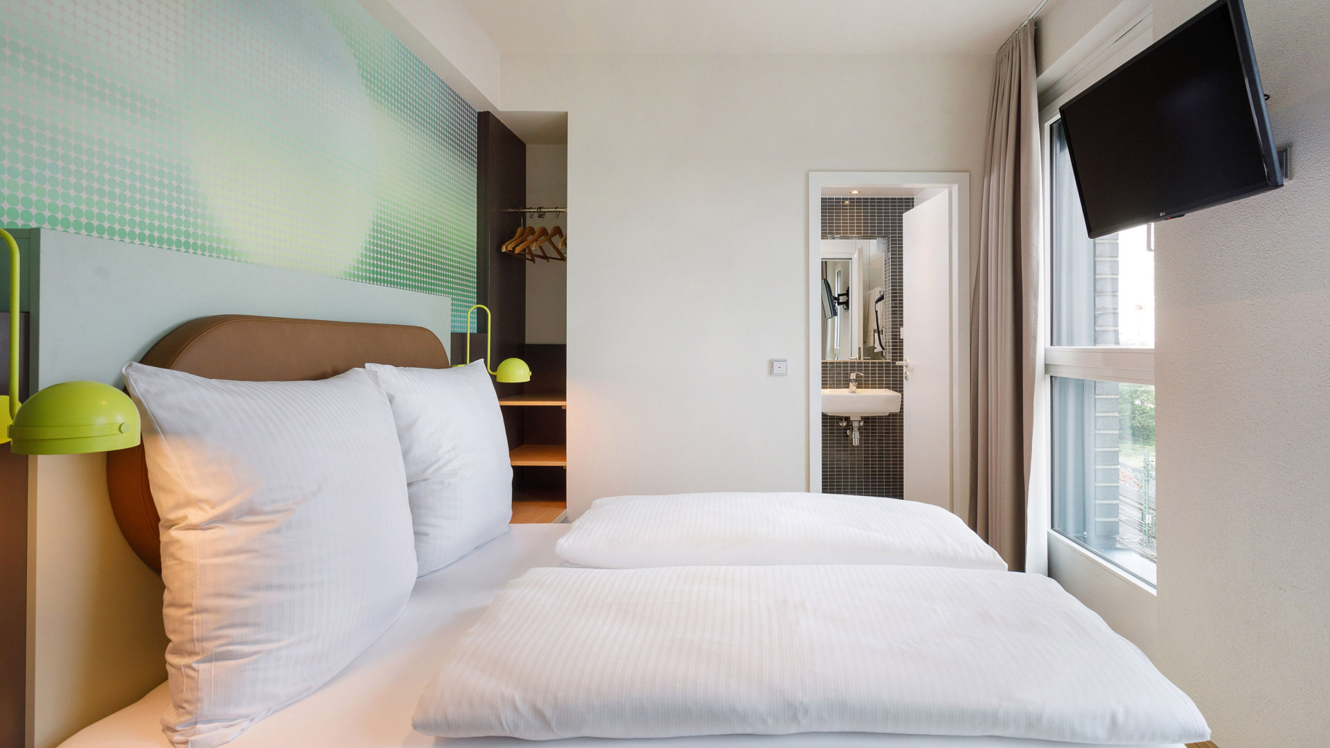 Großes Hotelbett mit weißen Bezügen in einem hellen Hotelzimmer, neben dem Bett dekorative gelbe Leuchten und Ferneseher.